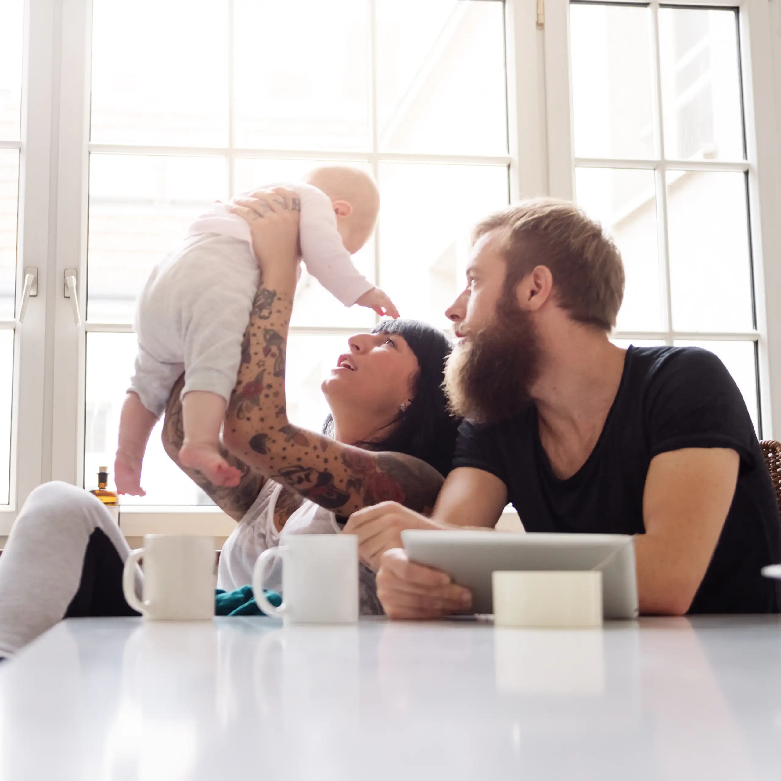 A young couple sit in front of a window holding a newborn baby