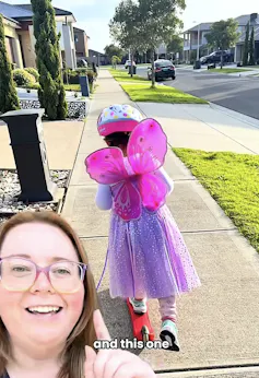 A small girl in fairy wings, a tutu and a helmet on a scooter, shown from behind, as her mum talks to the camera.