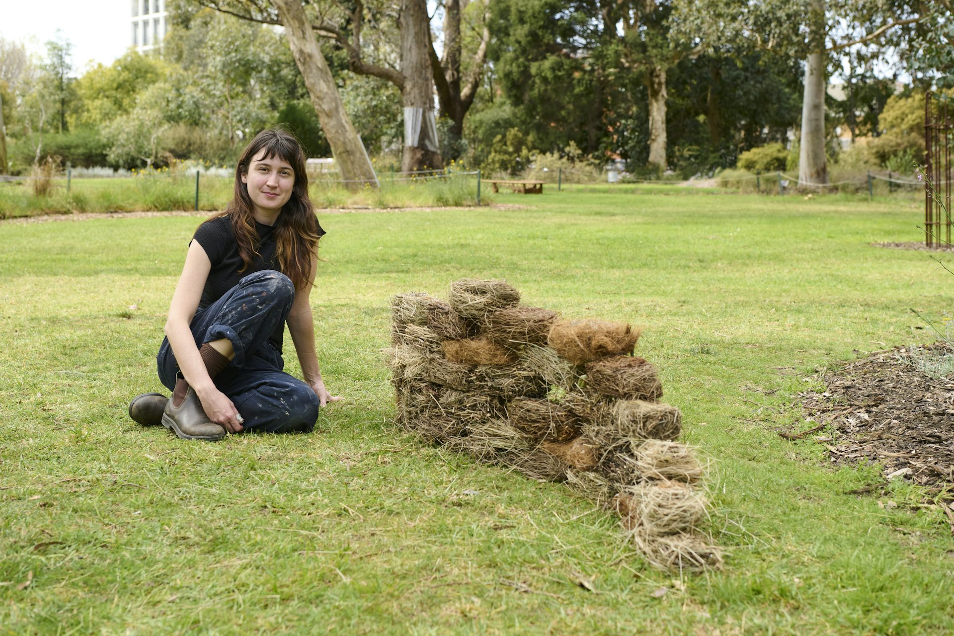 A young woman sitting on grass in a park beside a stacked wall of bricks made from natural materials.