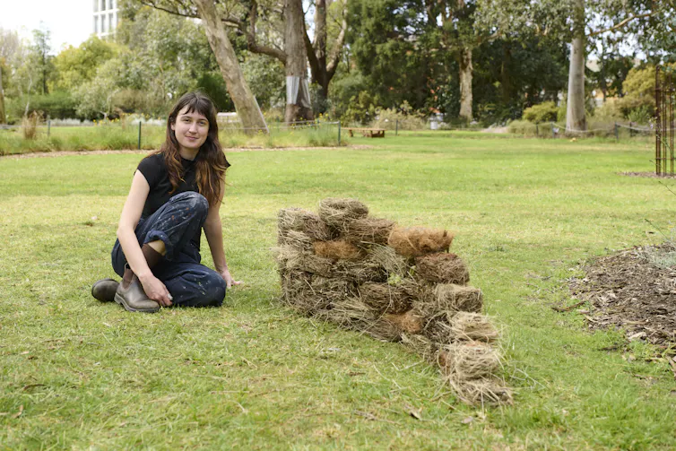 A young woman sitting on grass in a park beside a stacked wall of bricks made from natural materials.