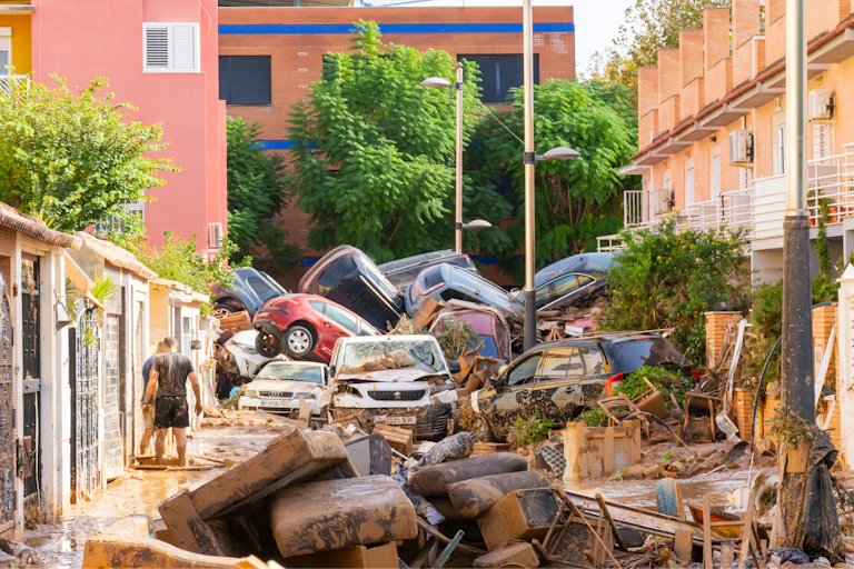 Dos persones en un carrer després de les inundacions, amb mobles, cadires i una muntanya de cotxes enmig dels edificis.