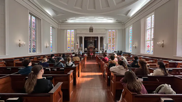 A religious sanctuary with stained-glass windows and young people sitting in wooden pews.