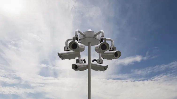 Several cameras are attached to a light post as clouds and blue sky appear in the background.