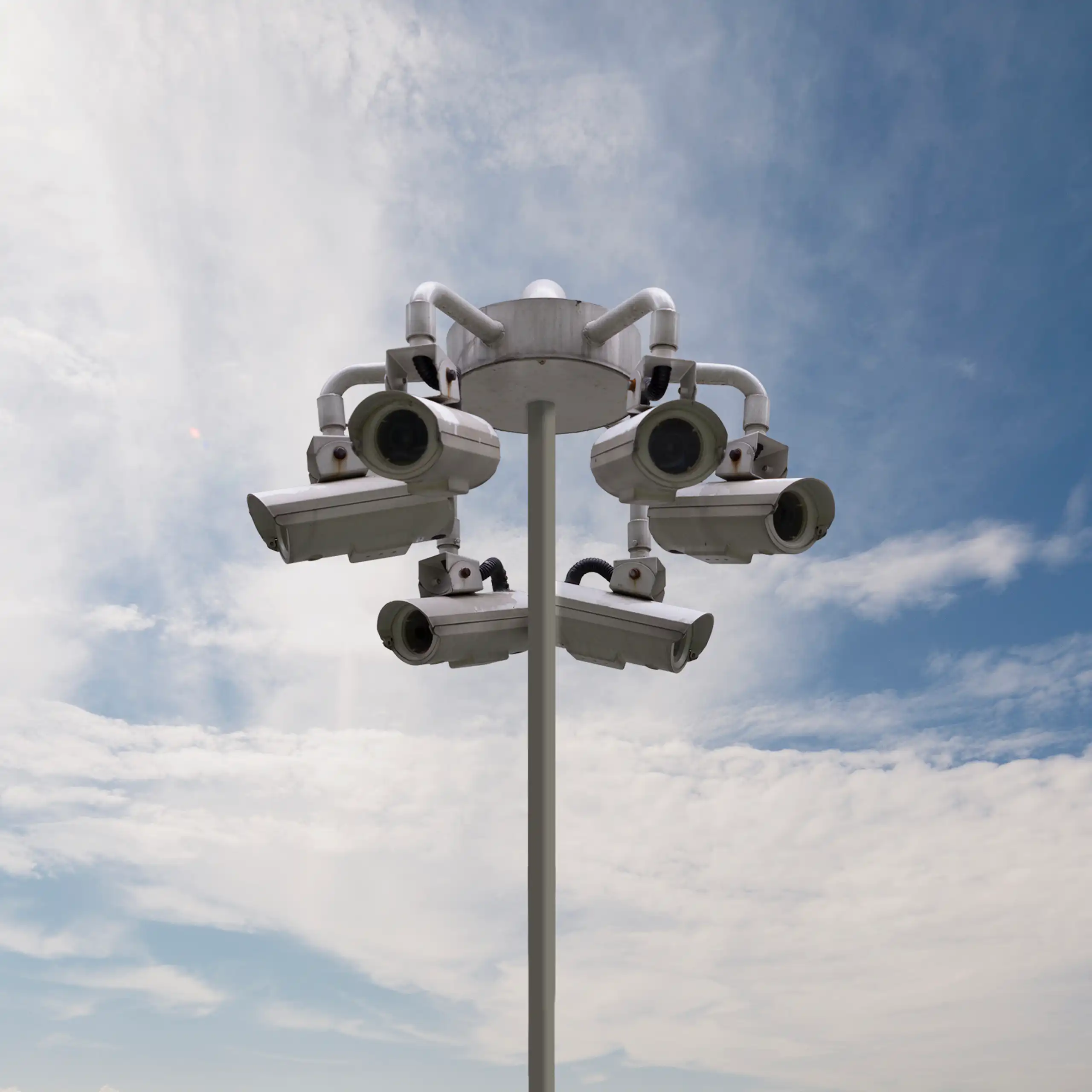 Several cameras are attached to a light post as clouds and blue sky appear in the background.