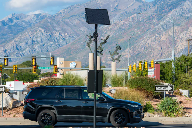 A black car drives by a post with a camera on it.