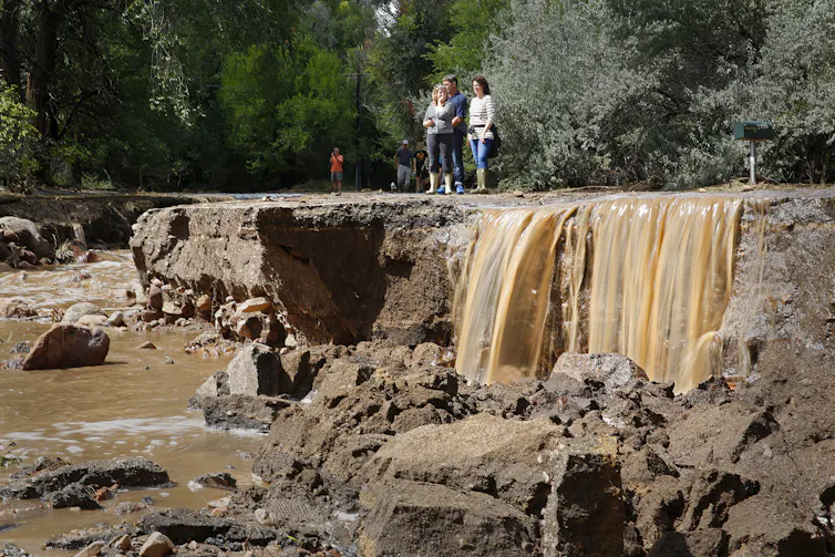 People stand on pavement looking at flowing water in a flood-damaged area.