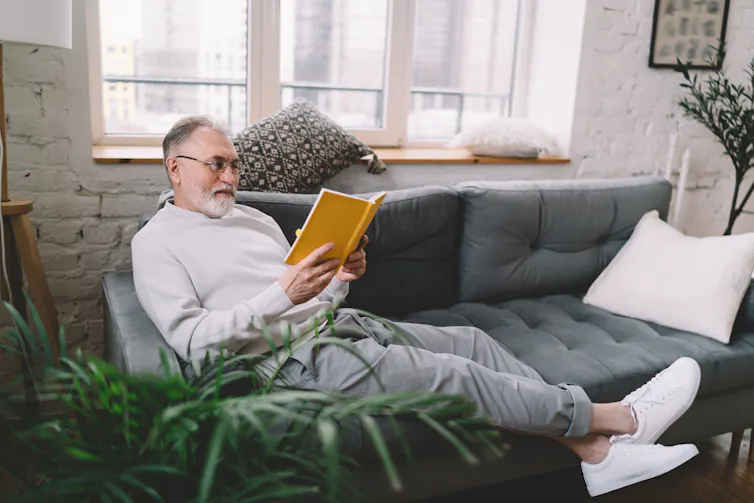 A man lazes on sofa reading a book and smiling with pleasure