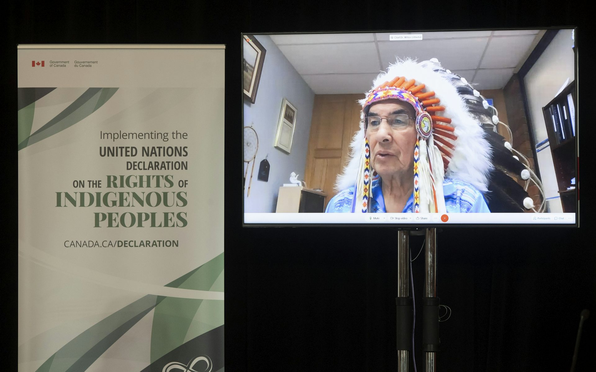 a man speaks on a screen wearing headdress feathered regalia in front of a sign about the UN Declaration on the Rights of Indigenous Peoples.