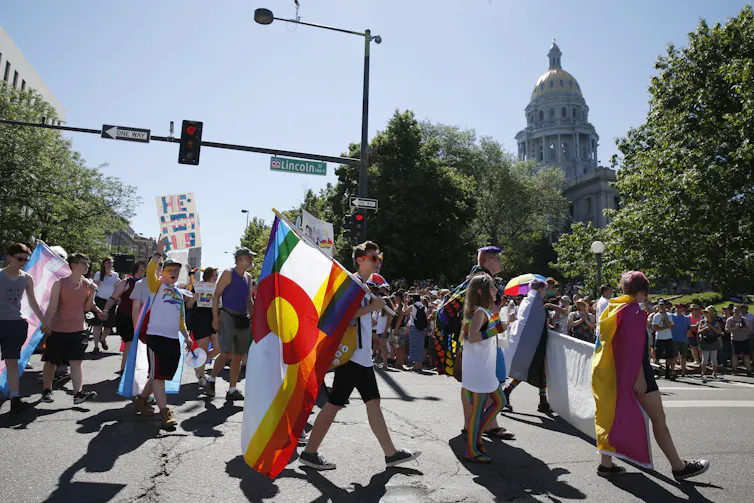 People in summer clothing, some of whom hold signs or brightly colored flags, walk down the middle of a broad street.