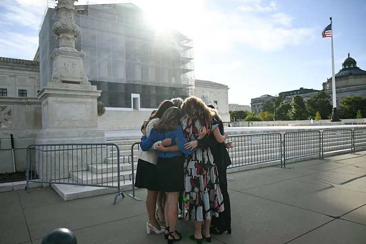 A small group of women huddle with their arms around each other outside a tall building with marble columns.