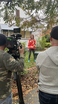 A woman stands in a garden while being filmed on camera.