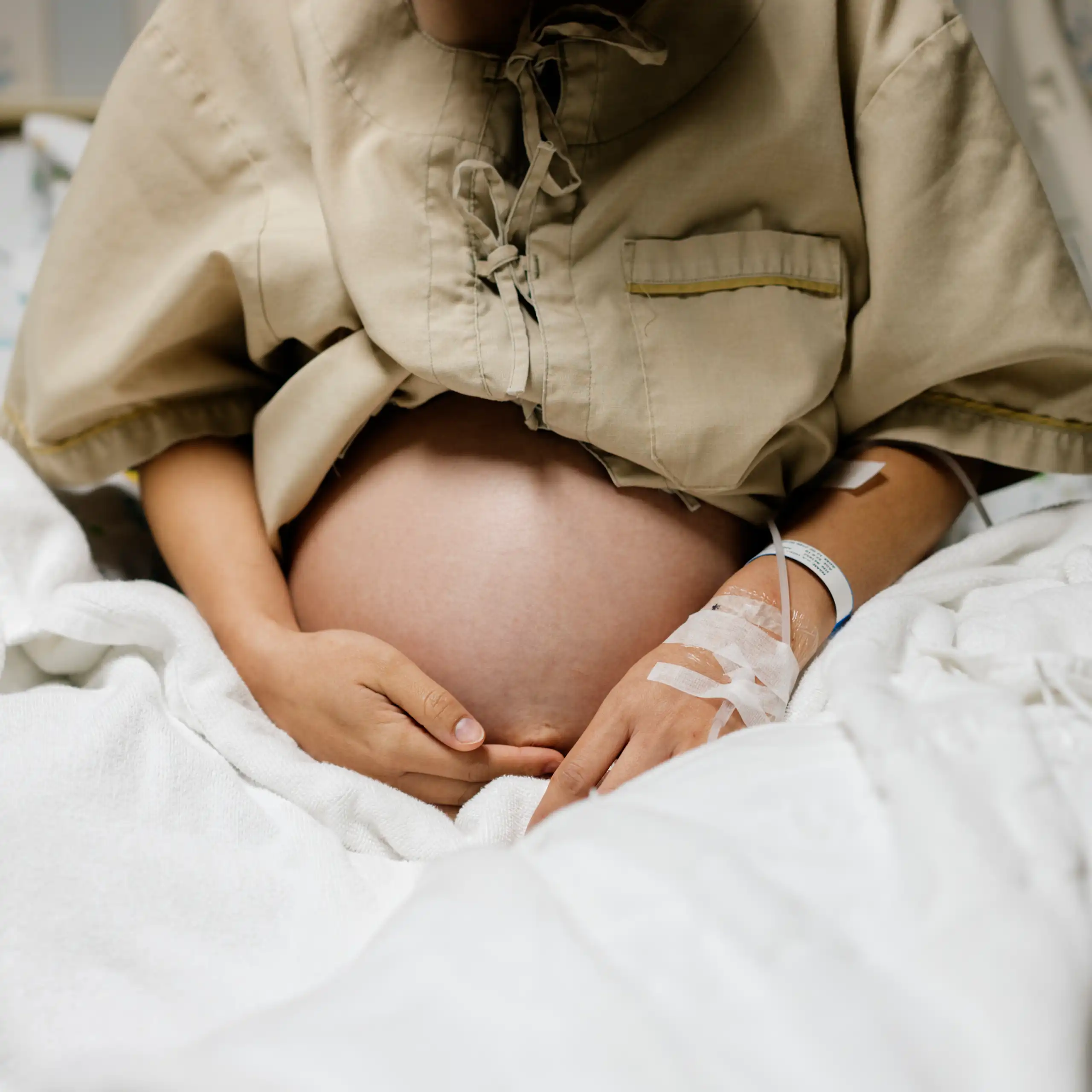 A pregnant person in a hospital gown and wristband, with an IV taped to one hand, rests in a hospital bed.