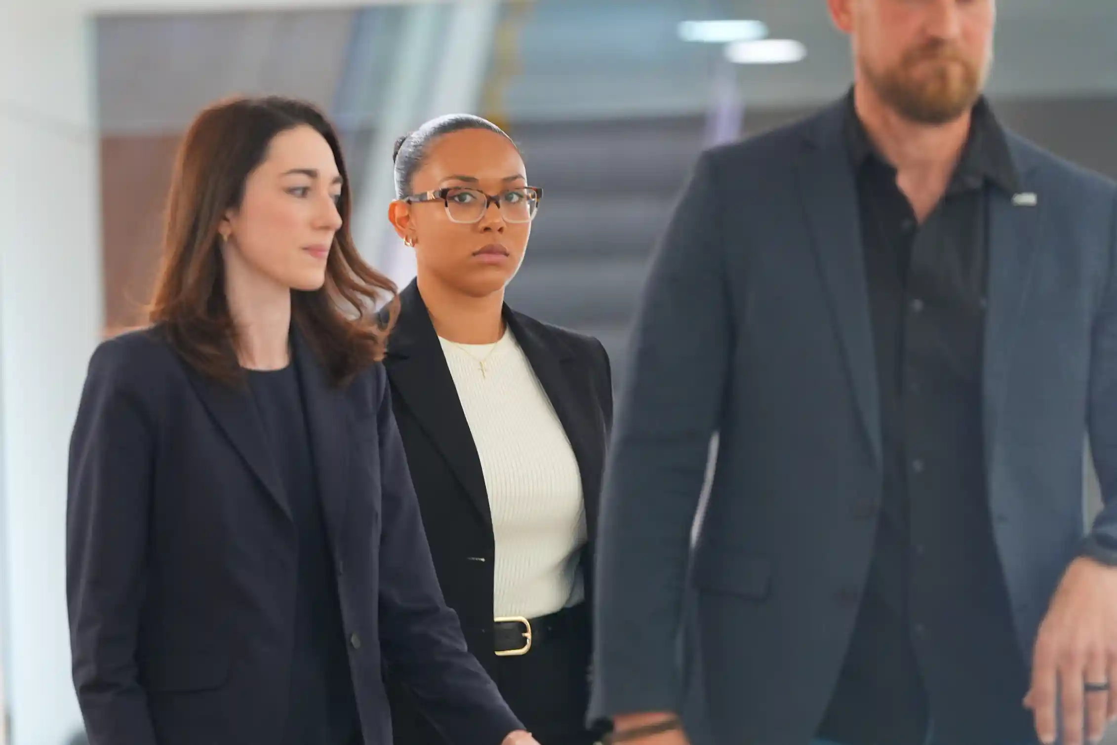 Two women and a man in dark business suits walk down a hallway