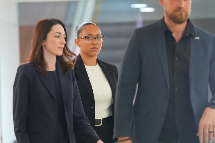 Two women and a man in dark business suits walk down a hallway