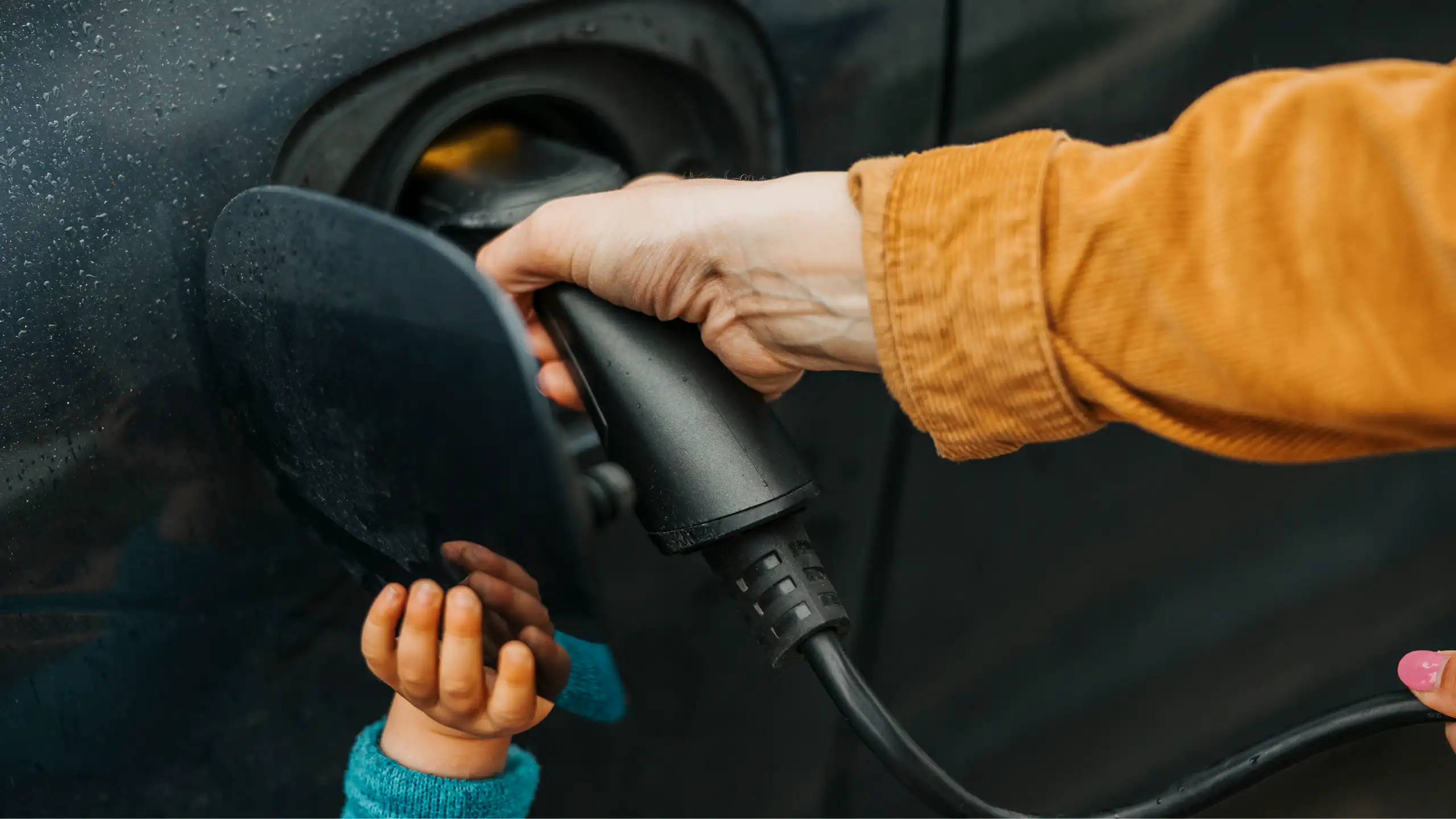 A woman's hand plugs a charger into a port, helped by a child's hand.