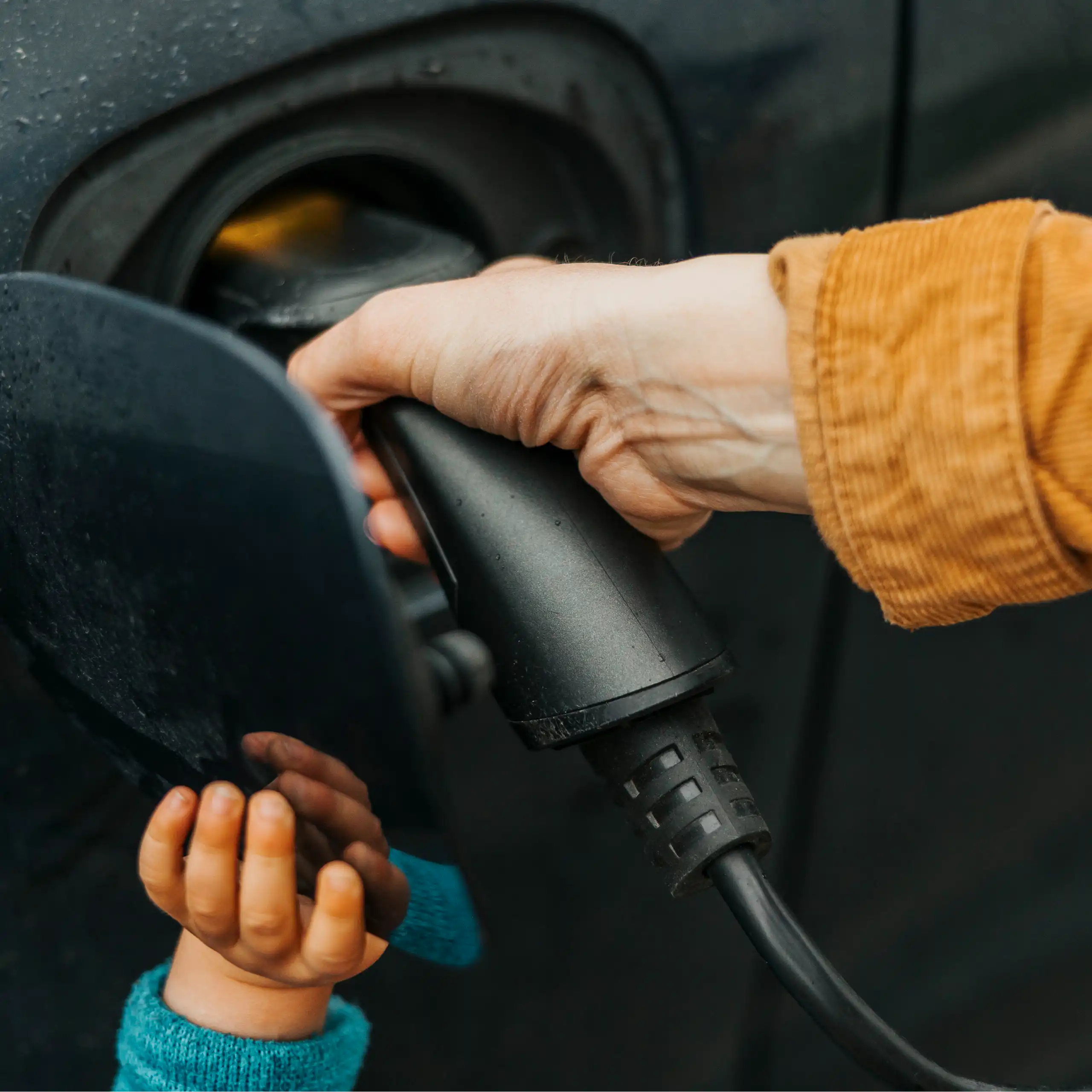 A woman's hand plugs a charger into a port, helped by a child's hand.