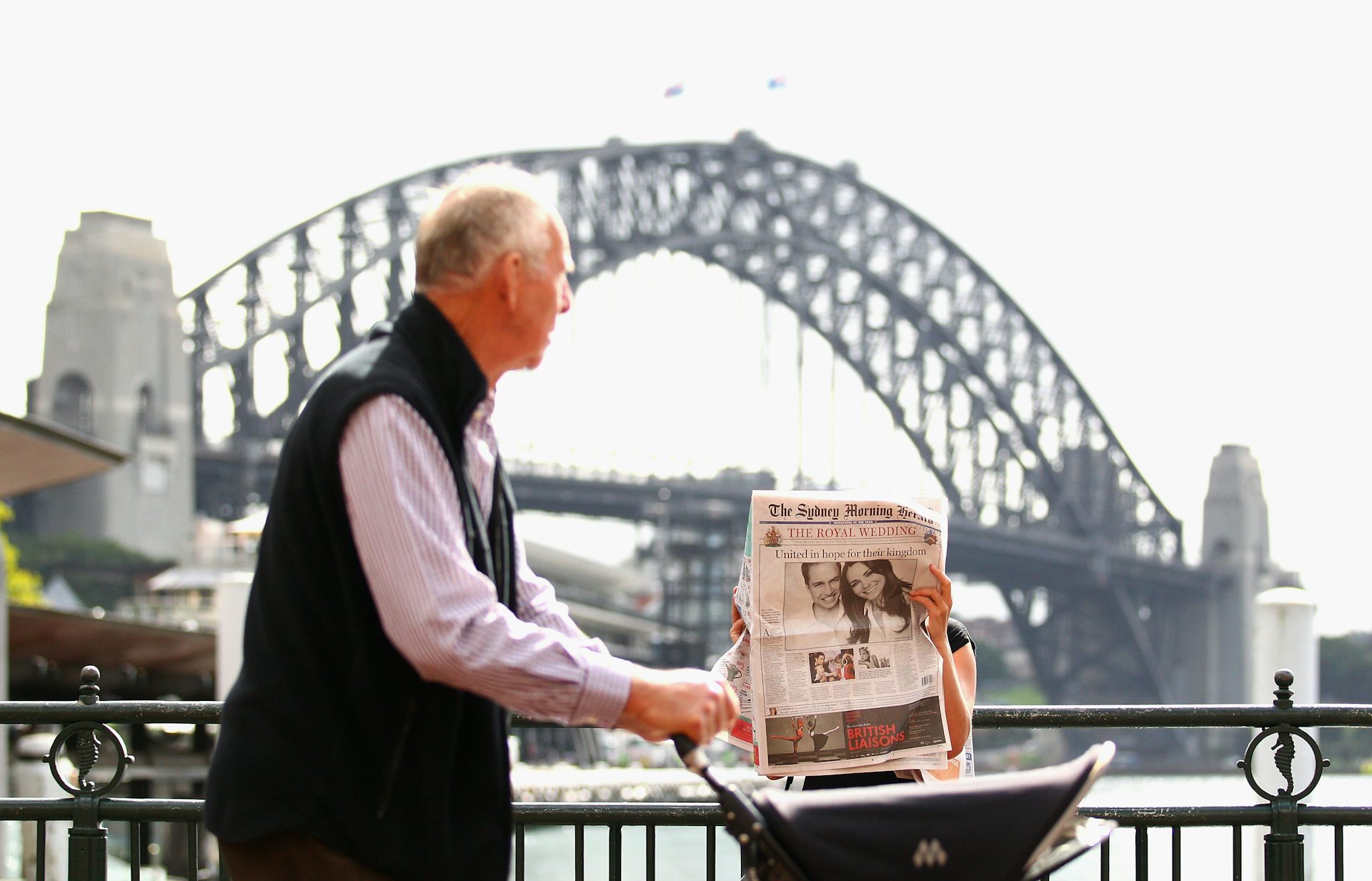 A grey-haired man pushes a pram along a path in front of the Sydney Harbour Bridge, while a woman in the background reads a newspaper.