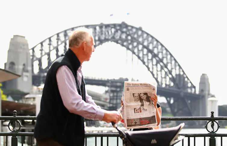 A grey-haired man pushes a pram along a path in front of the Sydney Harbour Bridge, while a woman in the background reads a newspaper.