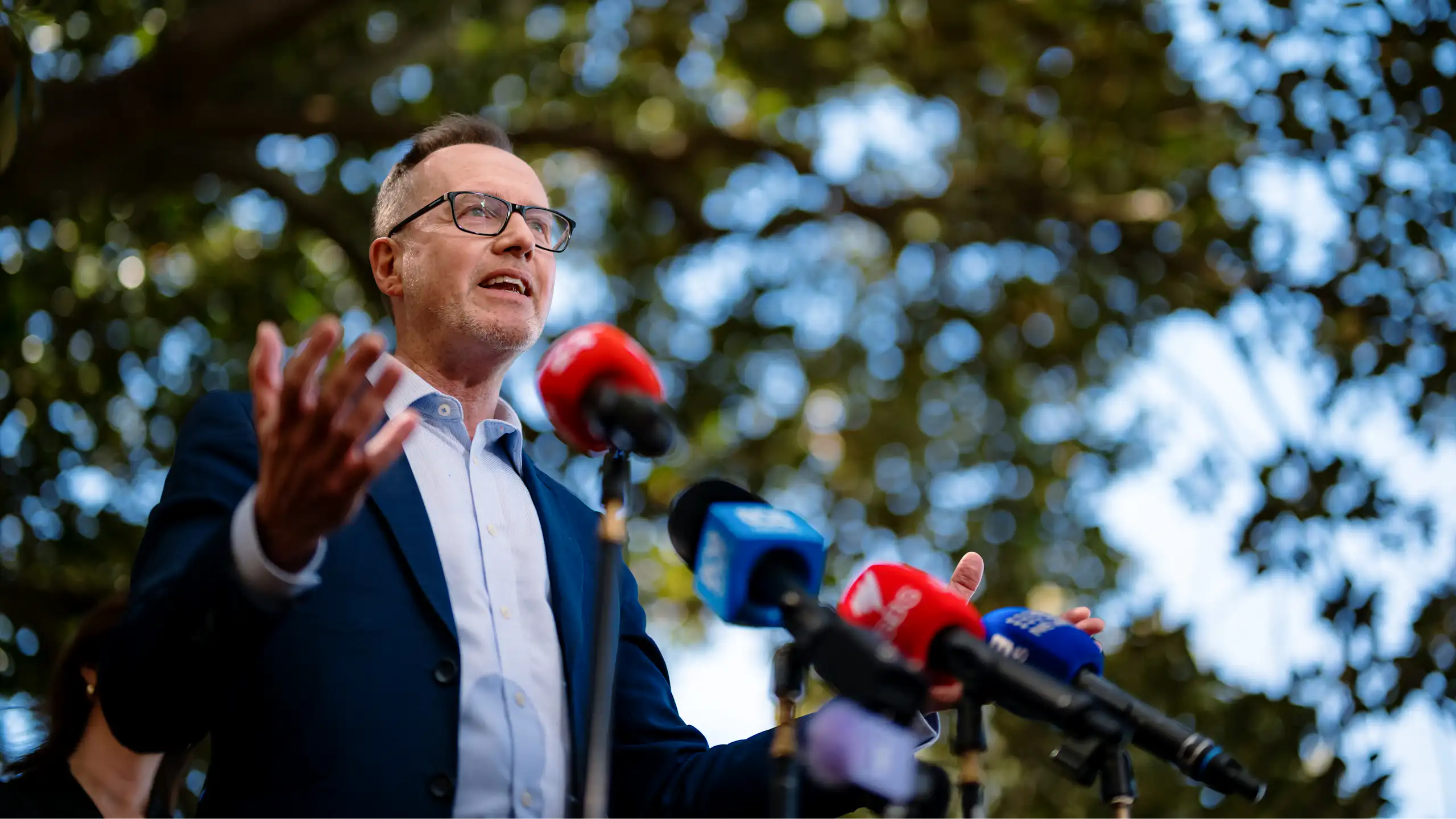 A man with glasses speaks to the media under a tree