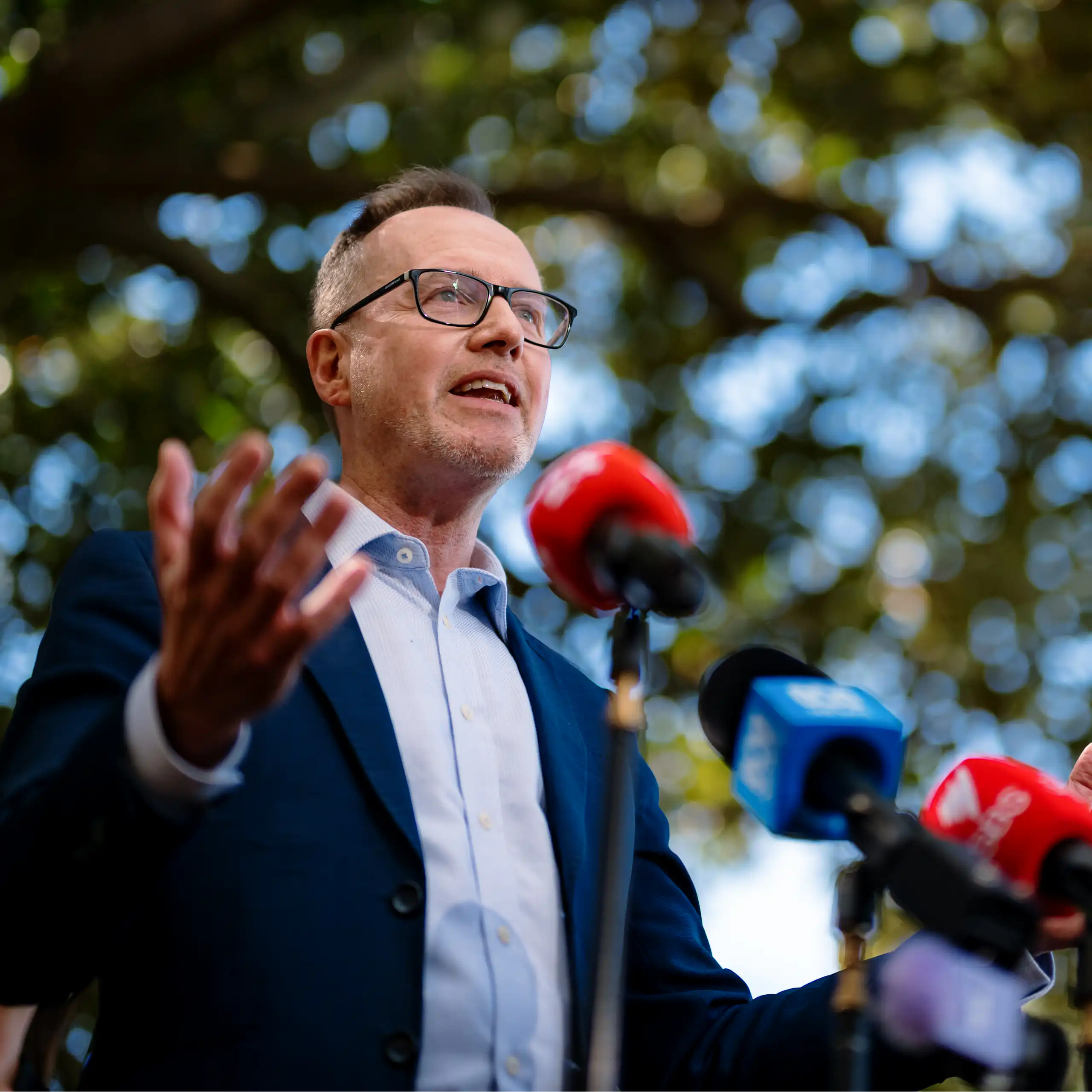 A man with glasses speaks to the media under a tree