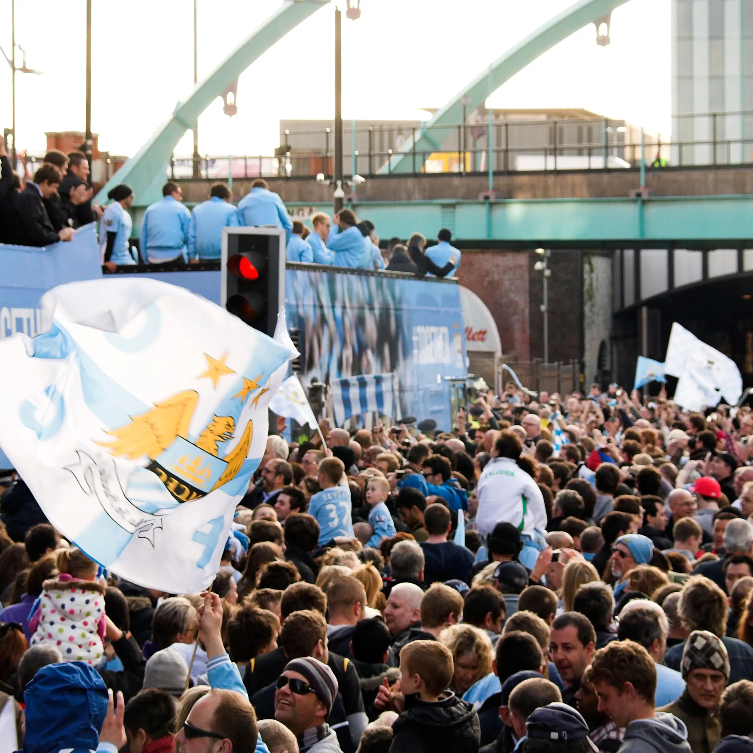 Photograph of crowded street, blue buses and Manchester City fans waving flags