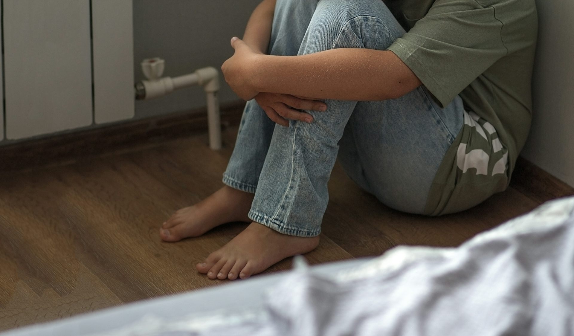 Une personne seule, une jeune femme ou un enfant, assise par terre repliée sur elle-même dans une chambre.