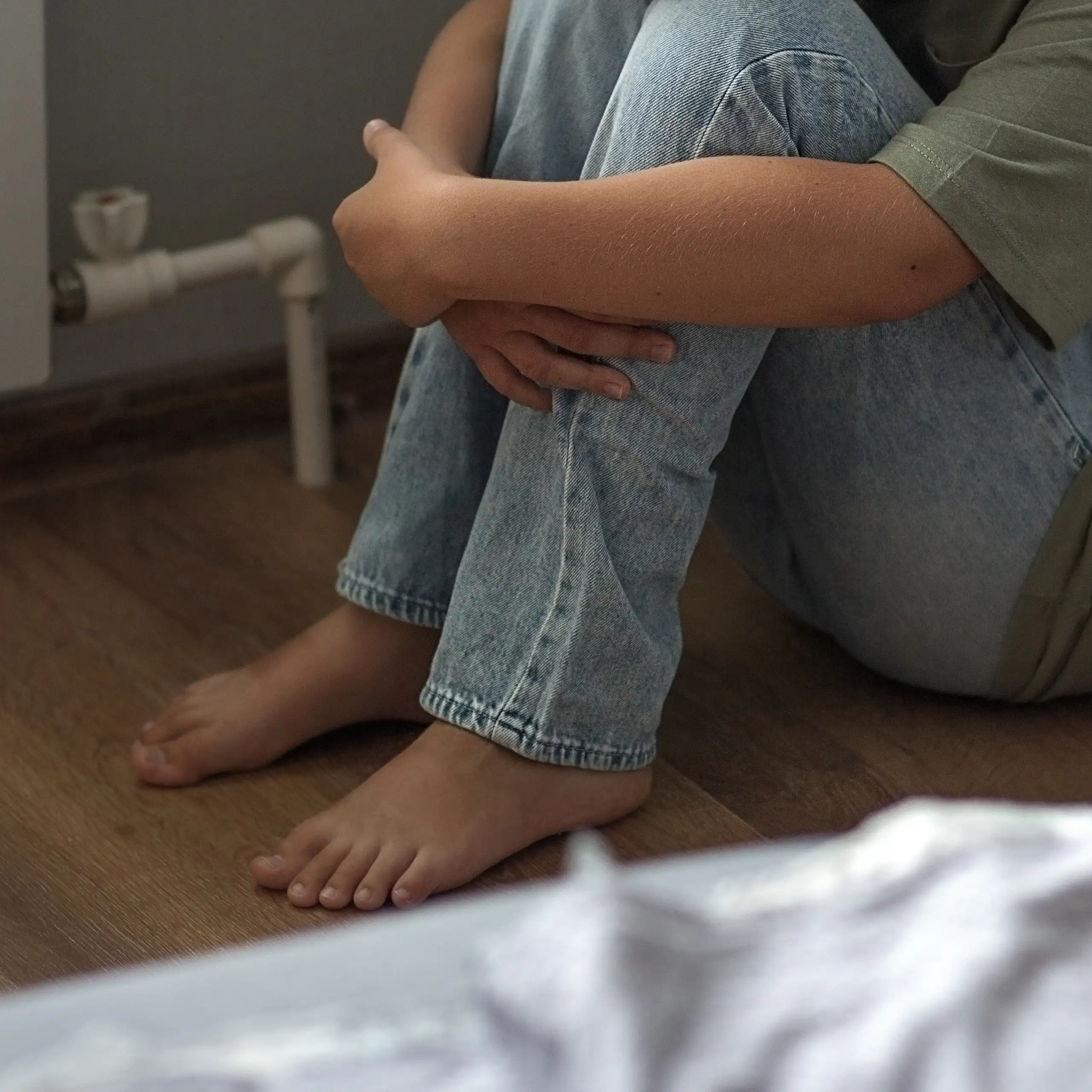Une personne seule, une jeune femme ou un enfant, assise par terre repliée sur elle-même dans une chambre.