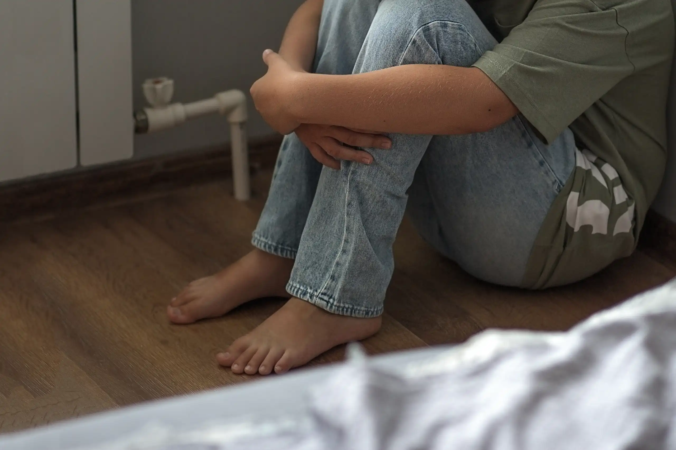Une personne seule, une jeune femme ou un enfant, assise par terre repliée sur elle-même dans une chambre.