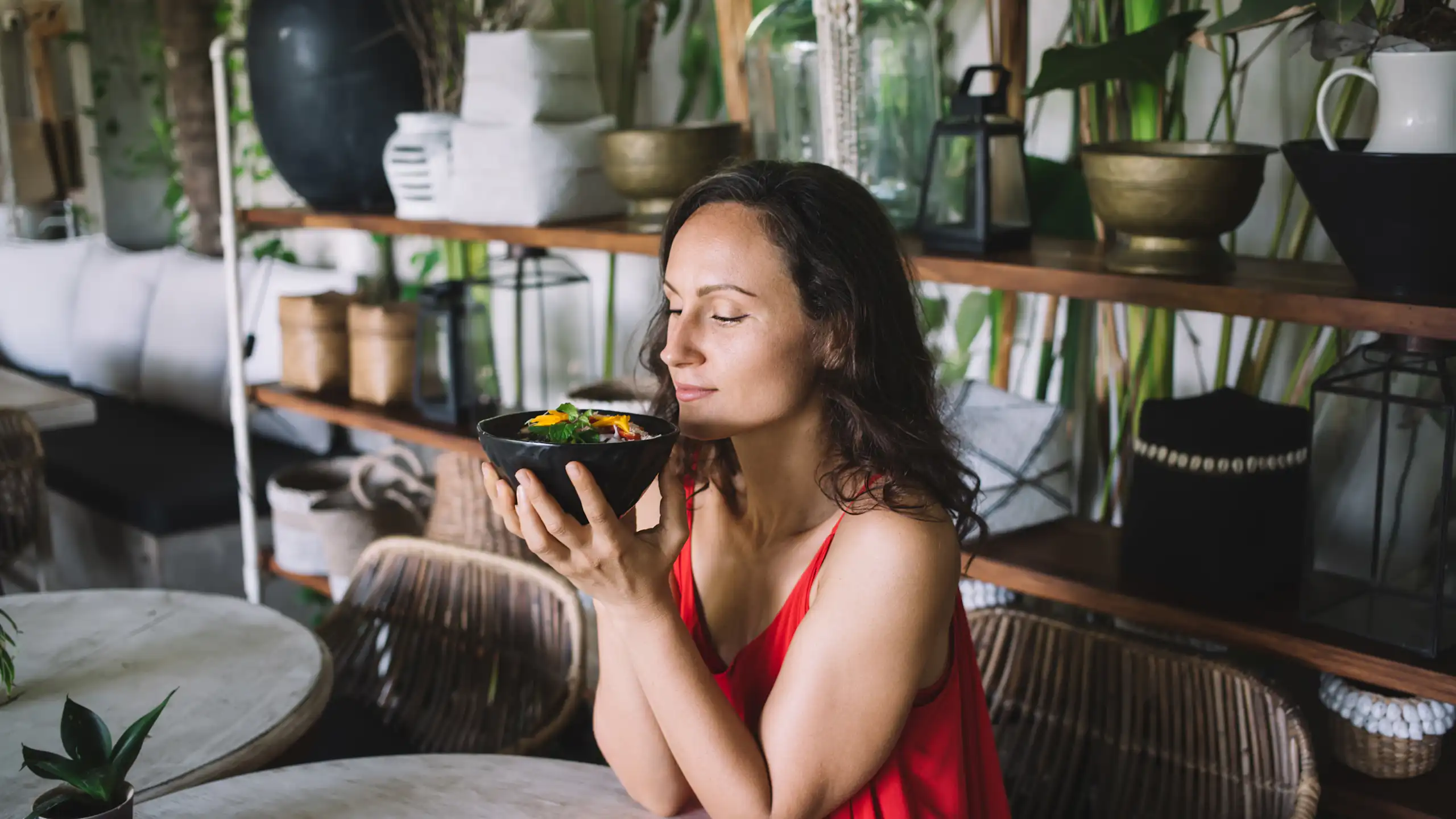 Entourée de plantes luxuriantes, une femme en robe rouge hume l'arôme d'un bol rempli de nourriture, les yeux fermés, et apprécie ce qu'elle sent.
