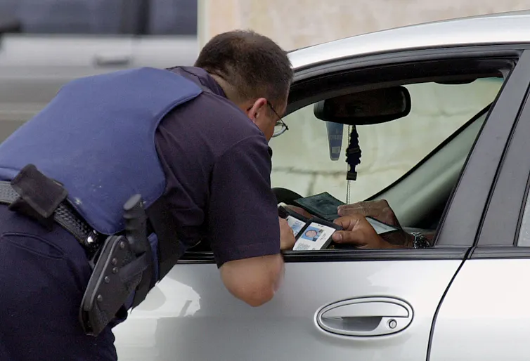 law enforcement officer leans into driver side of car looking at the ID the driver is holding out
