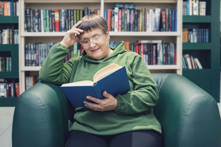 An elderly woman reads a book while sitting in a library.