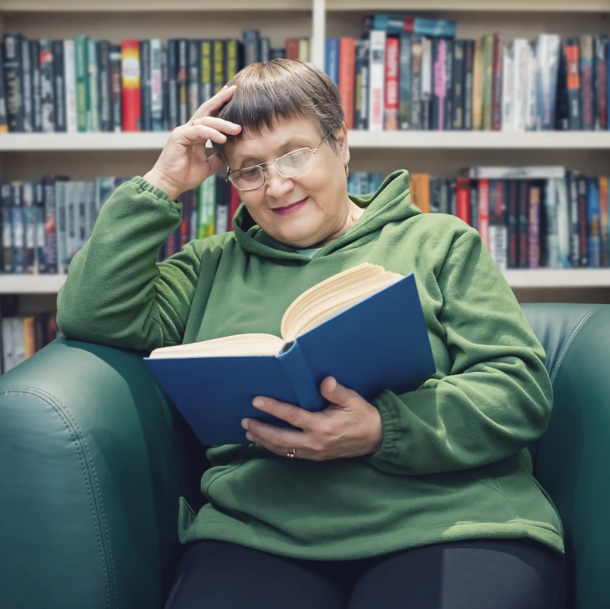 An elderly woman reads a book while sitting in a library.