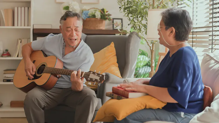 An elderly man plays an acoustic guitar while an elderly woman sings along.