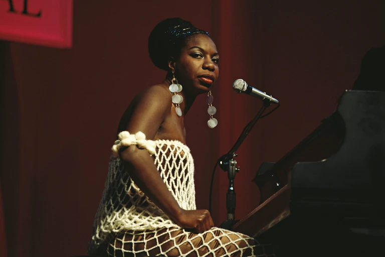 Young Black woman wearing a white, fishnet dress and long, dangling, silver earrings sits in front of a piano on a stage and looks out into the crowd.