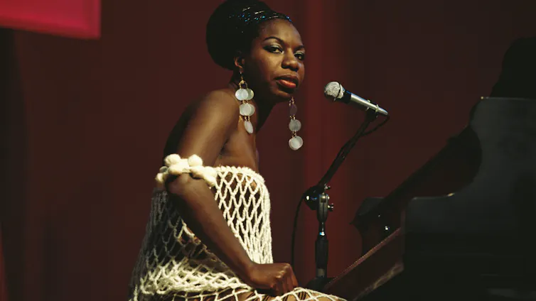 Young Black woman wearing a white, fishnet dress and long, dangling, silver earrings sits in front of a piano on a stage and looks out into the crowd.