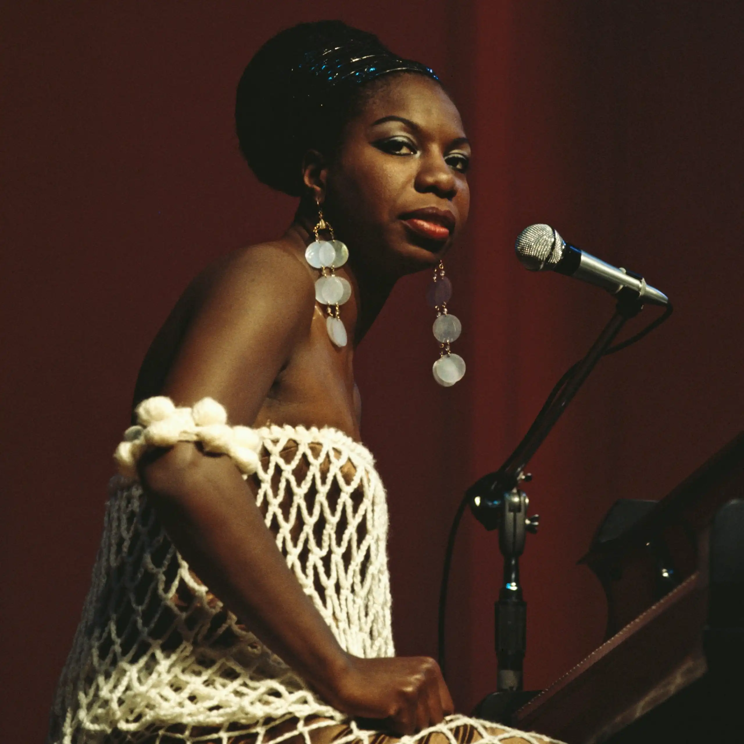 Young Black woman wearing a white, fishnet dress and long, dangling, silver earrings sits in front of a piano on a stage and looks out into the crowd.