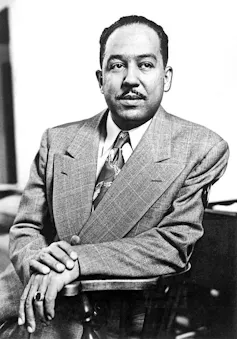 Middle-aged mixed-race man with a thin mustache posing while wearing a suit and sitting in a wooden chair.