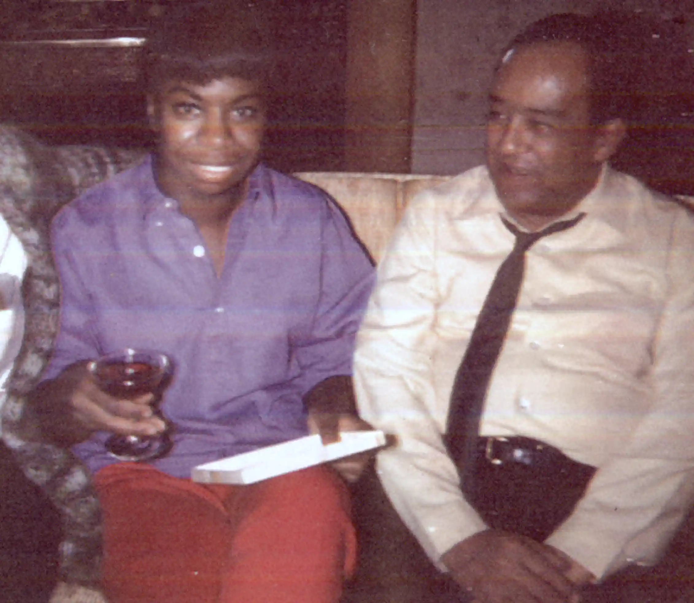 Young smiling Black woman holds a glass of wine and wears a loose purple shirt seated next to older Black man wearing a white dress shirt and tie.