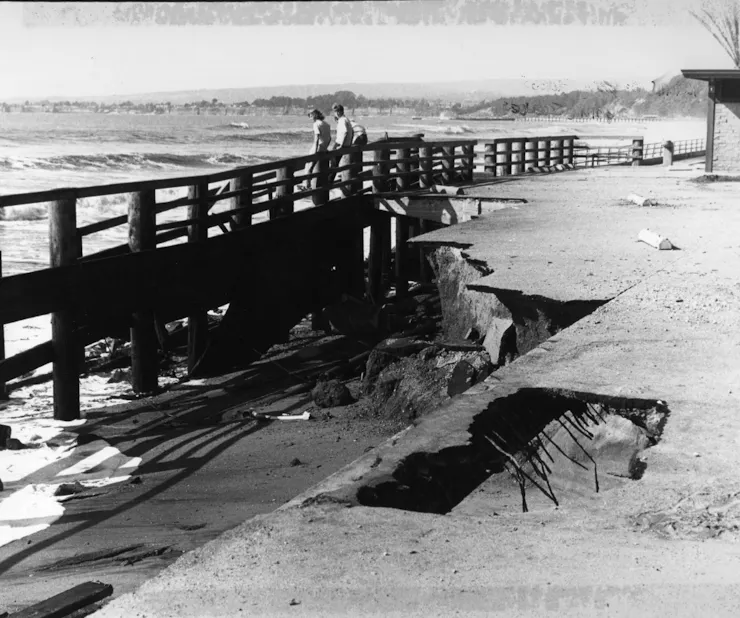 Two people stand on a remains of a damaged pier. Large slabs of the deck have fallen into the water.