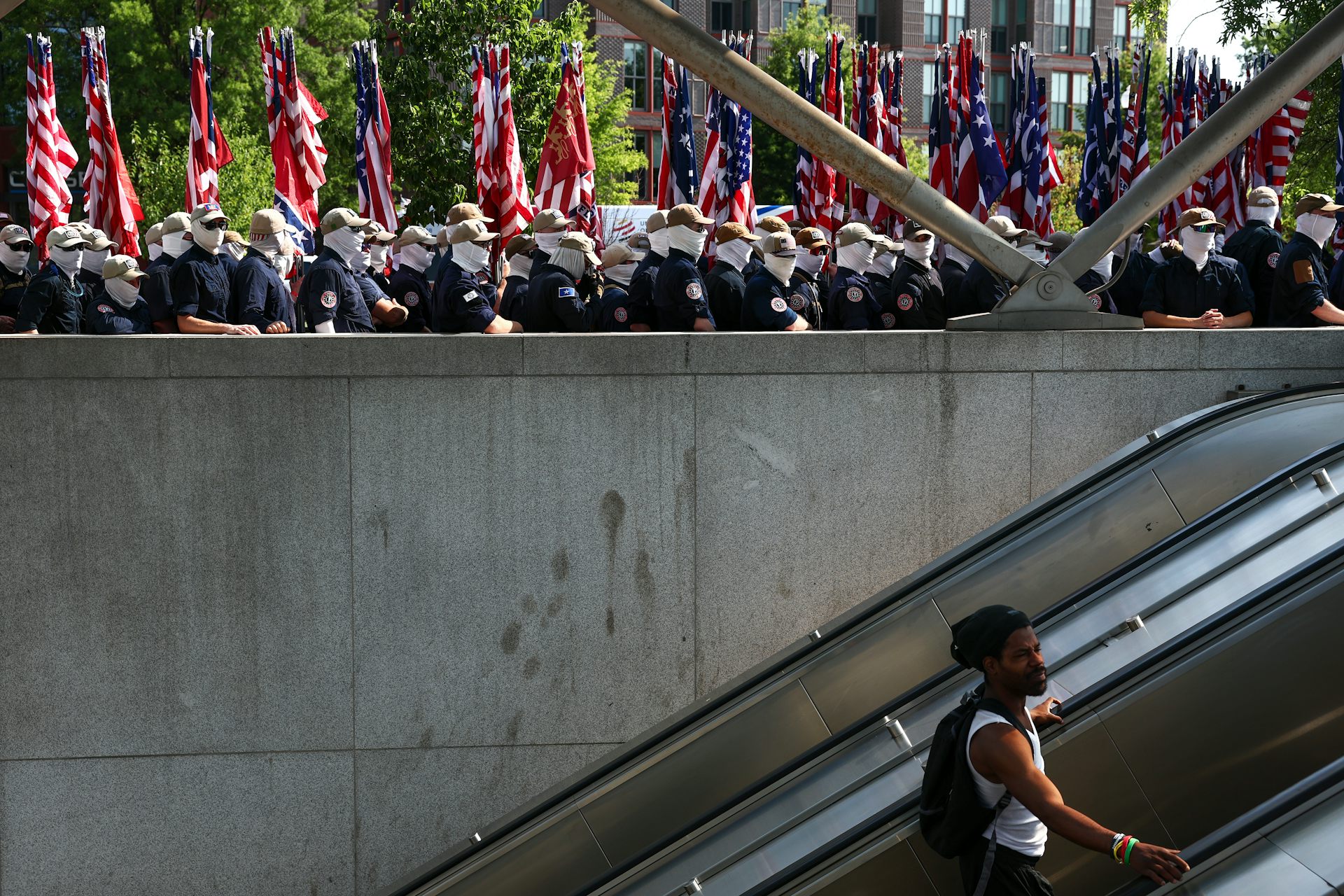 Men in white face masks and dark shirts stand at a railing above a subway escalator with American flags behind them as a man rides the escalator below