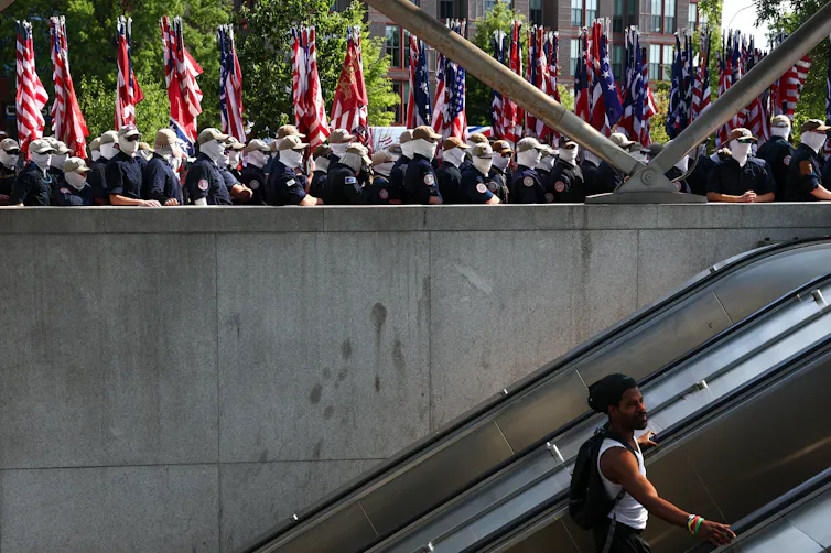 Men in white face masks and dark shirts stand at a railing above a subway escalator with American flags behind them as a man rides the escalator below