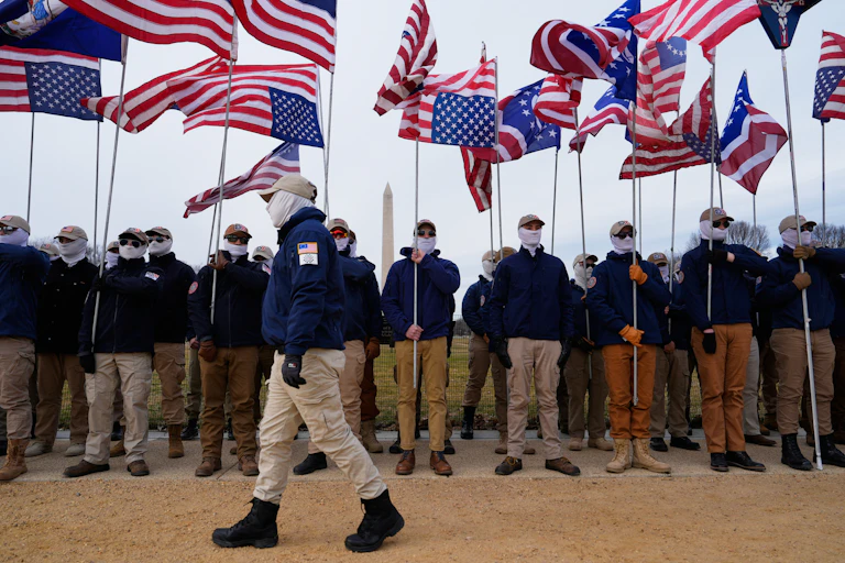 Men in white face coverings, dark jackets and khaki pants stand in a line holding flags in front of the Washington Monument as another man walks past.