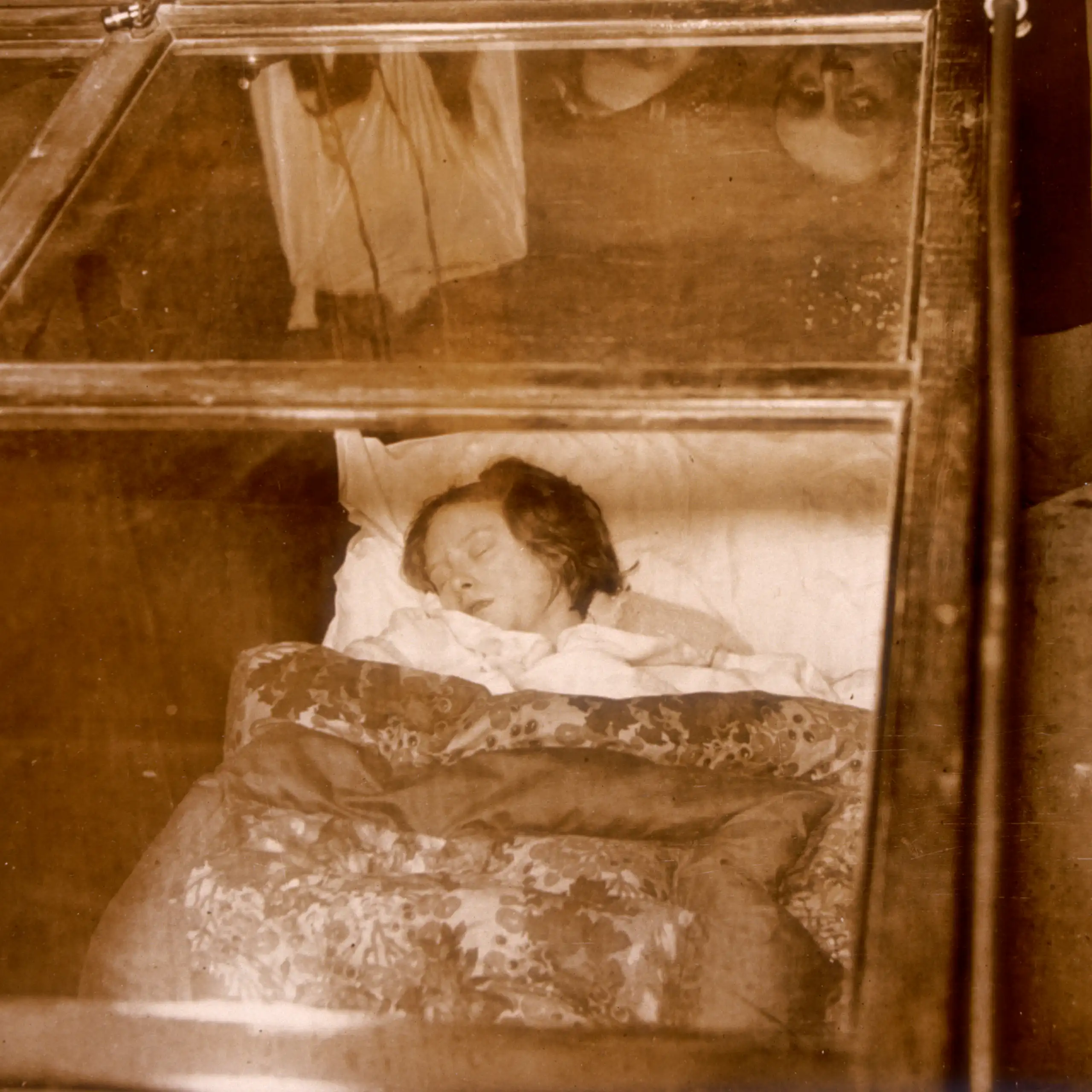 A woman sleeps in a glass box while people watch greedily.