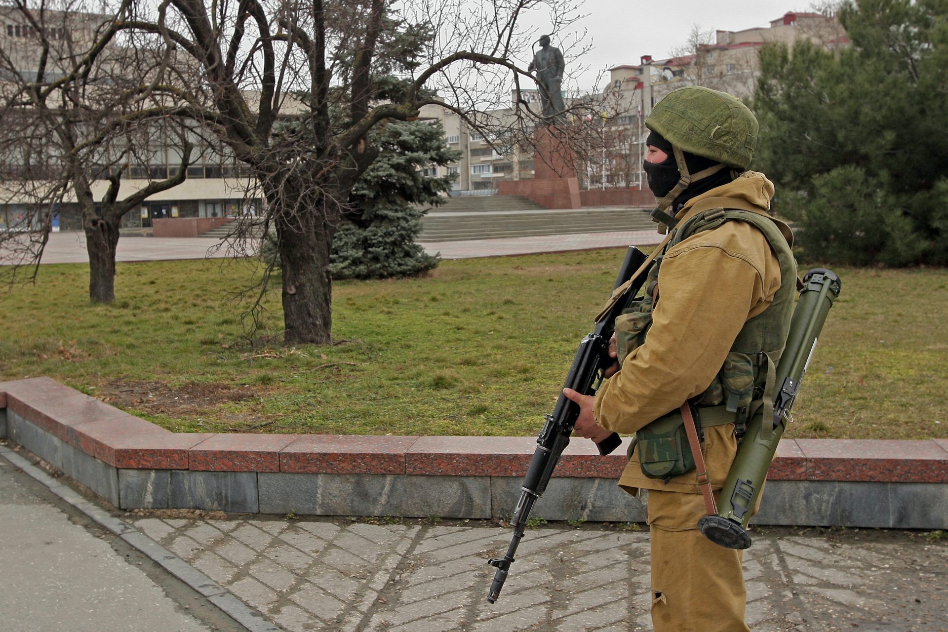 A man in military uniform and a face mask stands in parkland holding a rifle.