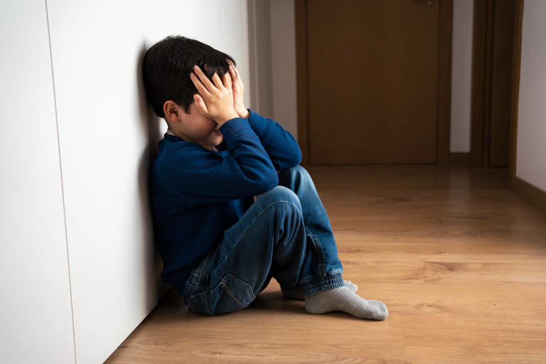 Niño con las manos en la cara sentado y apoyado contra una pared