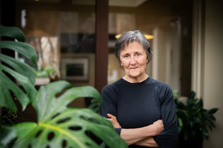 An older woman standing, arms crossed, by a monstera plant.