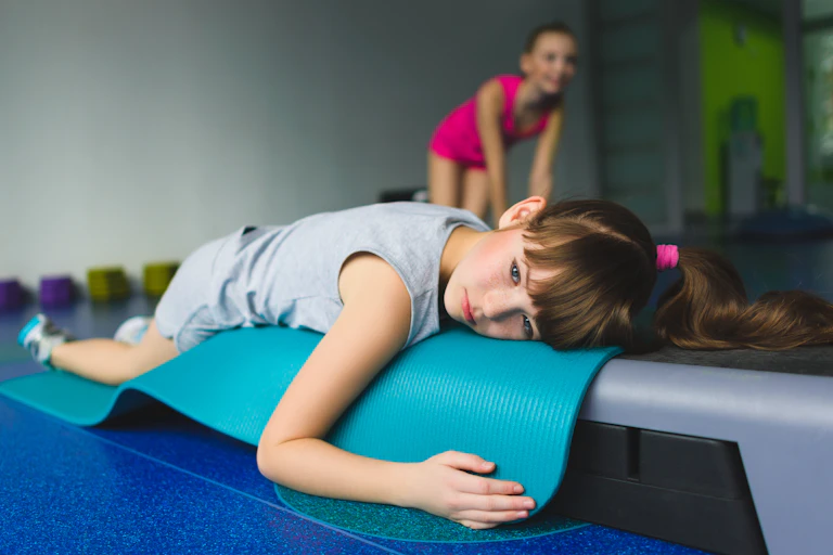 Tired girl lying on a fitness mat in PE class while classmate smiles and exercises in the background