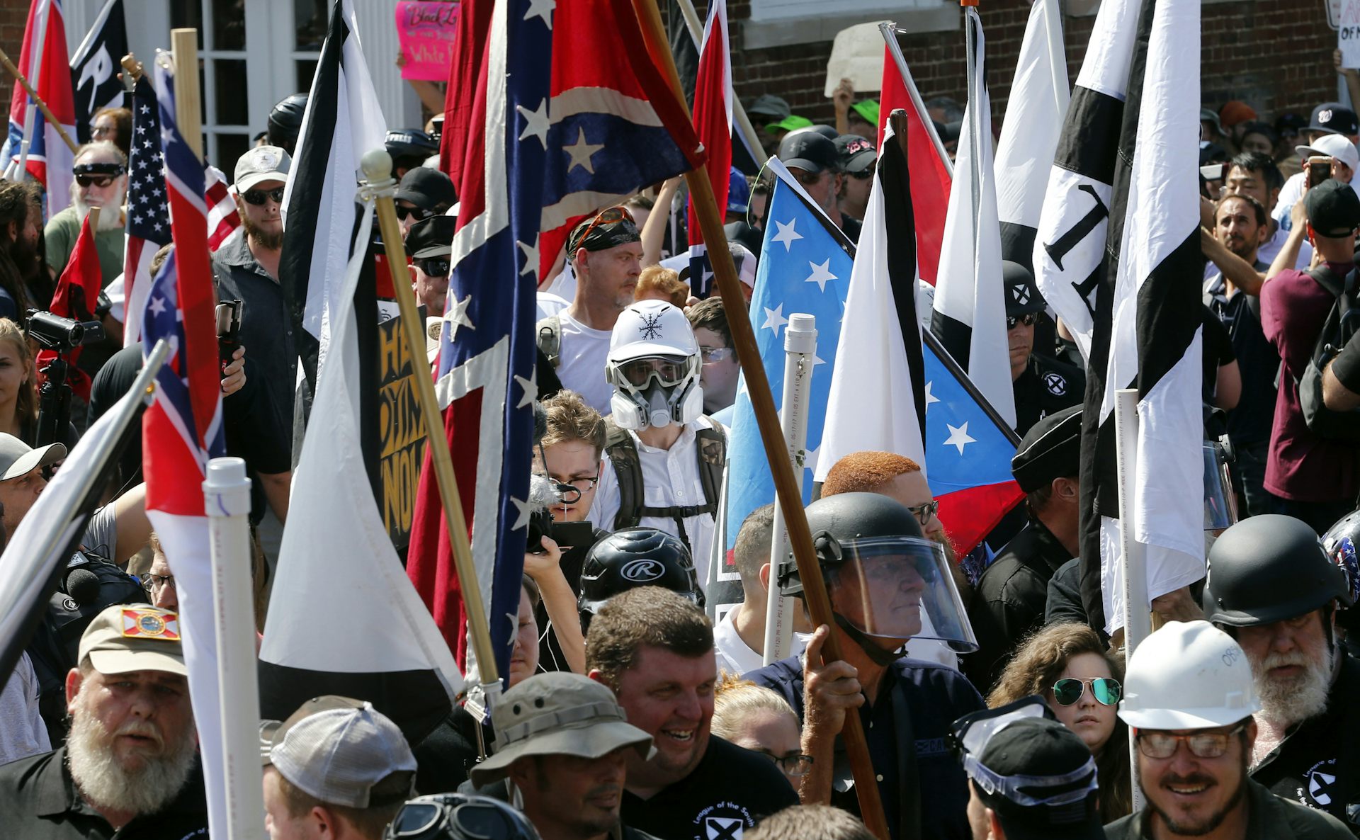 A crowd of men, some in helmets and protective gear, march with Confederate flags and other banners.