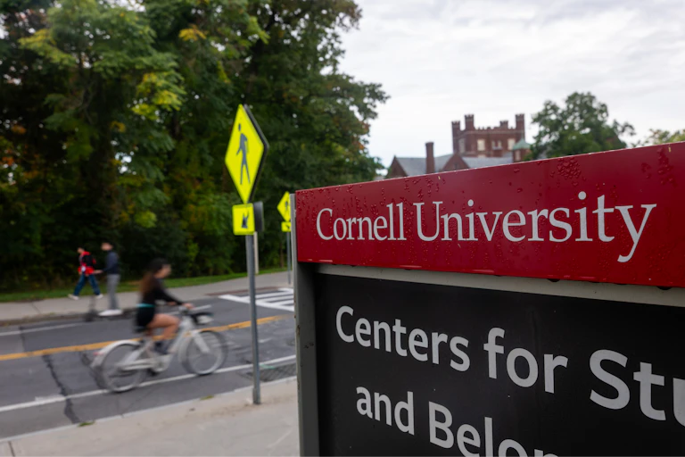 A person rides a bike down a quiet street in front of a red sign that says in white lettering 'Cornell University.'
