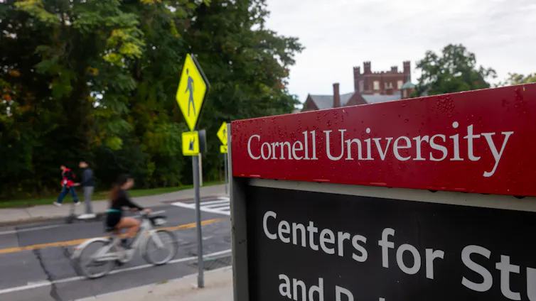 A person rides a bike down a quiet street in front of a red sign that says in white lettering 'Cornell University.'